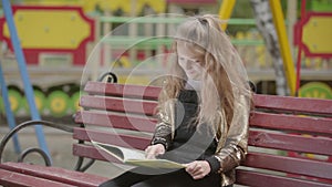Cute teen girl is reading a book while sitting on a bench outdoors.