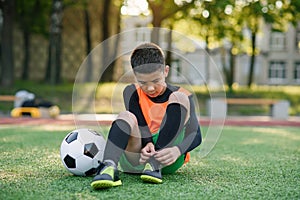 Cute teen soccer player on artificial green covering of outdoors sport field tying the shoelace on boots after training.