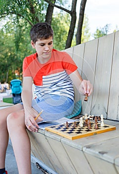 Cute Teenage boy plays chess on the bench.  Trendy caucasian teen in white t shirt sitting in the city park with wooden chess.