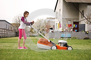 Cute teenage child girl working in garden, mowing green grass with lawn-mower on backyard on summer holiday. Concept of