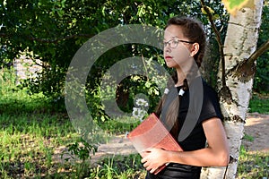 cute teenage girl with glasses is leaning against tree in summer park and reading book. Ð¡oncept of training