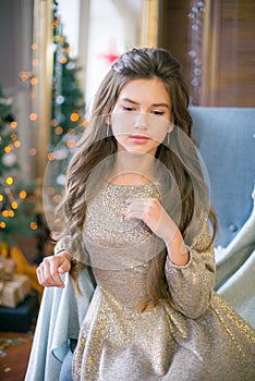 A cute teenage girl with long curly hair in a shining dress in a room decorated for Christmas with shining garlands