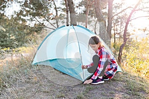 A cute teenage girl pitches a tent in the middle of a pine forest. The concept of summer active recreation, travel and adventure,