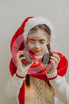 A cute teenage girl in a pullover and Santa Claus hat holds and eats a chocolate cookie against the background of a light wall.