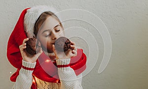 A cute teenage girl in a pullover and Santa Claus hat holds and eats a chocolate cookie against the background of a light wall.