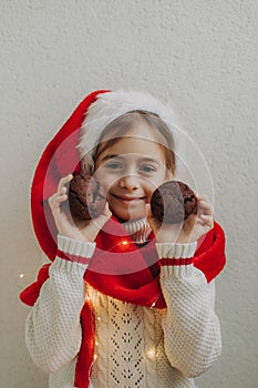 A cute teenage girl in a pullover and Santa Claus hat holds and eats a chocolate cookie against the background of a light wall.