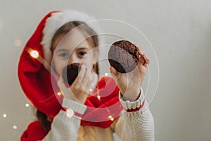 A cute teenage girl in a pullover and Santa Claus hat holds and eats a chocolate cookie against the background of a light wall.