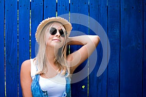 Cute teenage hipster girl smiling in straw hat and sunglasses outdoor over blue wooden background.