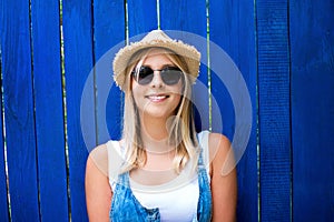 Cute teenage hipster girl with toothy smile in straw hat and sunglasses outdoor over blue wooden background.