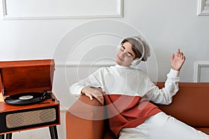 Cute teenager boy wearing earphones listening music and having fun, chilling on sofa near wooden turntable with vinyl record