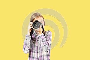 Cute teenager girl posing with digital SLR photo camera, huge zoom lens, strapped her neck. Isolated yellow background.