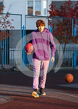 Cute teenager in violet hoodie playing basketball. Young boy with ball learning dribble and shooting on the city court. Hobby