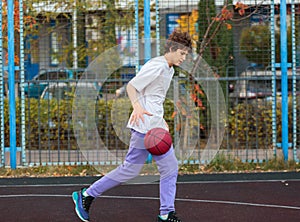 Cute teenager in a white t-shirt playing basketball outside. Young boy with ball learning dribble and shooting on the city court