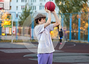 Cute teenager in a white t-shirt playing basketball outside. Young boy with ball learning dribble and shooting on the city court.