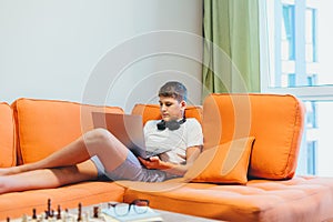 Cute teenager in white t shirt sitting on couch next to laptop and study. Serious boy in headphones makes homework,