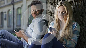 Cute teenagers sitting under tree, glancing at each other with loving looks