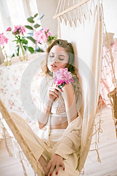 Cute tender young girl with blond long hair surrounded by pink peonies is resting in a hammock in the studio. Spring and flowers.