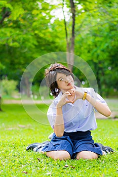 Cute Thai schoolgirl is sitting on the grass and doing heart symbol.