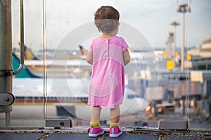 Cute Toddler baby in a pink dress looks at the planes at the airport. Waiting for a flight flight. back view