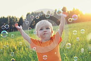 Cute toddler blond boy playing with soap bubbles on summer field. Happy child summertime concept. Authentic lifestyle image