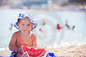 Cute toddler boy, blond child, eating watermelon on the beach coast