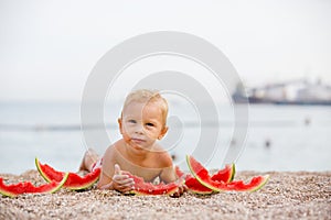 Cute toddler boy, blond child, eating watermelon on the beach coast