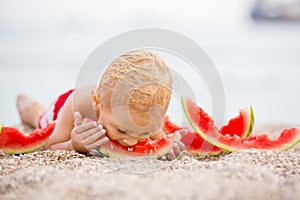 Cute toddler boy, blond child, eating watermelon on the beach coast
