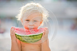 Cute toddler boy, blond child, eating watermelon on the beach coast