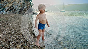 Cute toddler boy collecting pebbles on rocky beach and throwing them in sea water