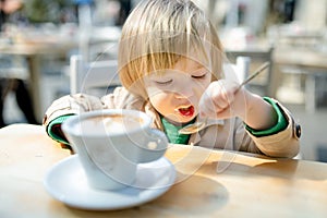 Cute toddler boy having hot chocolate in outdoor cafe. Small child drinking hot beverage on sunny terrace in Lecco. Spending good