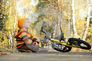 Cute toddler boy sitting on empty road in autumn park looking on falling orange leaves. Child`s bicycle lies nearby. Fall vibes