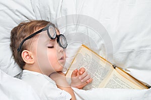 A cute toddler boy is sleeping in a round glasses with an old book under a white blanket.