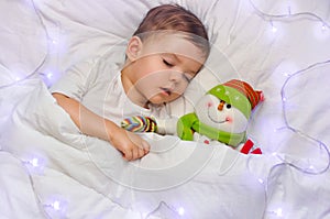 A cute toddler boy is sleeping on white linen with his favorite toy snowman in the blue lights of the garland.