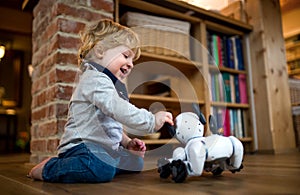 A cute toddler boy standing indoors at home, playing with robotic dog.
