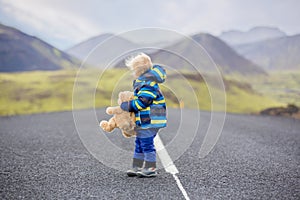 Cute toddler boy with teddy bear in hand, running on a road in Iceland