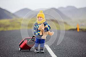 Cute toddler boy with teddy bear and suitcase in hand, running on a road in Iceland