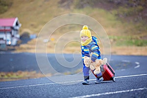 Cute toddler boy with teddy bear and suitcase in hand, running on a road in Iceland