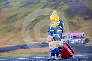 Cute toddler boy with teddy bear and suitcase in hand, running on a road in Iceland