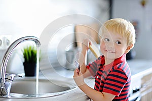 Cute toddler boy washing dishes in domestic kitchen