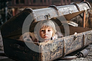 Cute toddler child, blond boy, hiding in a big suitcase in the attic, looking scared