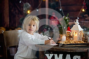 Cute toddler child, boy on Christmas, writing letter to Santa Claus and eating cookies at home, wooden cottage