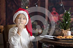 Cute toddler child, boy on Christmas, writing letter to Santa Claus and eating cookies at home, wooden cottage