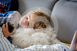 Cute toddler child, boy, lying on the couch, playing with mobile phone with his pet dog next