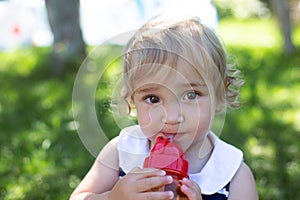 Cute toddler girl with big eyes drinking water from baby bottle outdoors in sunny summer day.