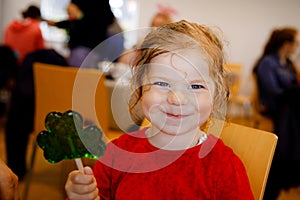 Cute toddler girl with cloverleaf lollipop in Irish pub or cafe. Child resting with parents and eating unhealthy sweets.