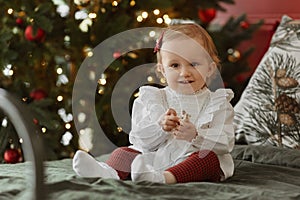 Cute toddler girl in dress sits on the bed with festive Christmas lights in the background