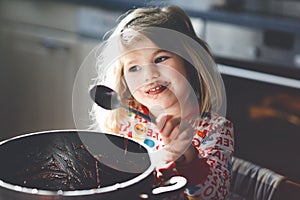 Cute toddler girl eating chocolate dough rests with spoon and fingers from pot. Happy child licking sweet dough for