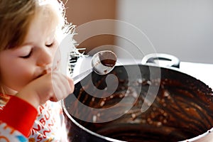 Cute toddler girl eating chocolate dough rests with spoon and fingers from pot. Happy child licking sweet dough for