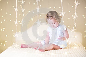 Cute toddler girl with her toy bear on a white bed between beautiful warm Christmas lights