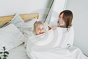 Cute toddler girl long fair hair big grey eyes looking at camera having fun with mother on bed